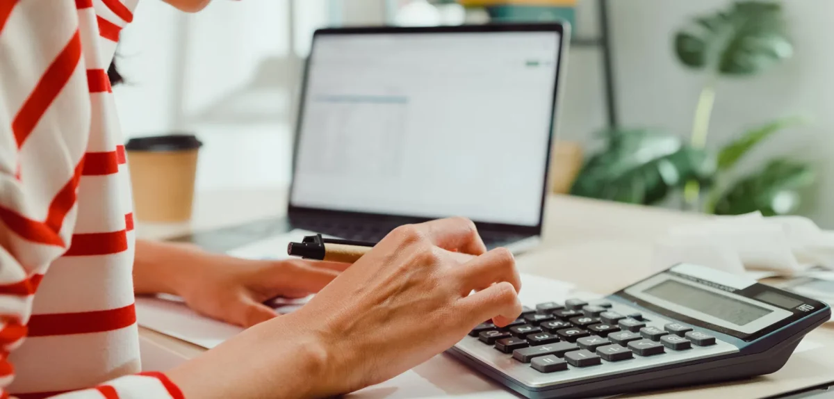 A person in a striped shirt calculates on a calculator while using a laptop, with a coffee cup nearby.