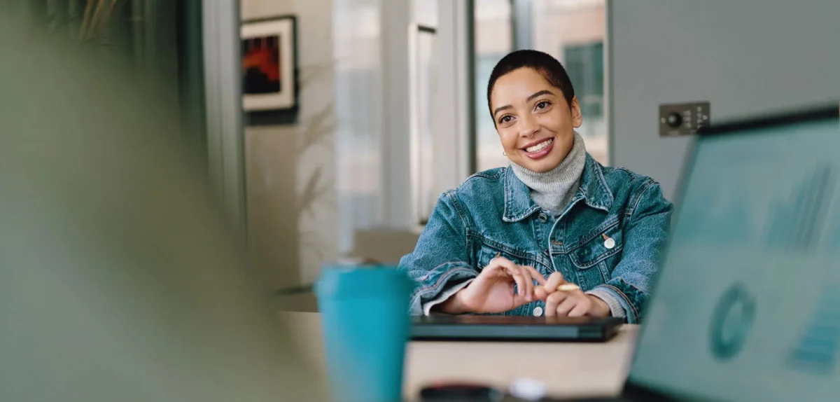 A person in a denim jacket sits at a table in a modern office, hands resting on a laptop, with a teal cup nearby and graphs on a screen.